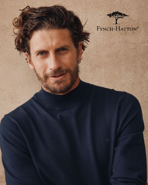 Un hombre con cabello castaño ondulado y barba lleva un jersey azul marino y está de pie frente a una pared beige con textura. El logo y el nombre de Fynch-Hatton aparecen en la esquina superior derecha.