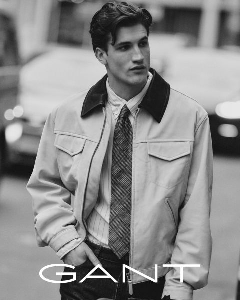 Un hombre que lleva una camisa con cuello, una corbata y una chaqueta clara sale al exterior con las manos en los bolsillos. La imagen está en blanco y negro, con la inscripción "GANT" en la parte inferior.