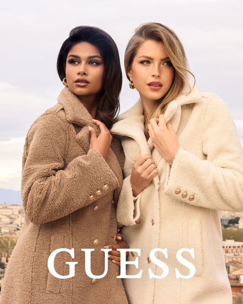 Dos mujeres están de pie juntas al aire libre, vistiendo abrigos texturizados, una en color canela claro y otra en color crema. Ambas sostienen sus cuellos y miran hacia adelante. La palabra "GUESS" está en negrita en letras blancas en la parte inferior.