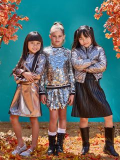 Tres jóvenes están de pie con confianza entre hojas de otoño, vistiendo atuendos brillantes y metálicos. Dos chicas llevan chaquetas plateadas, y la chica del centro lleva un top y una falda con lentejuelas. Un fondo verde azulado contrasta con el follaje naranja del otoño.