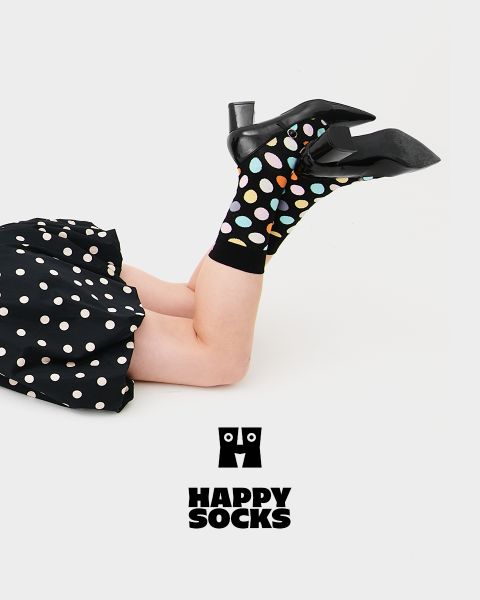 Una persona con tacones negros y calcetines negros con lunares de colores está acostada de lado, con las piernas en alto, sobre un fondo blanco. Lleva una falda negra de lunares. Debajo aparece el logotipo de Happy Socks.