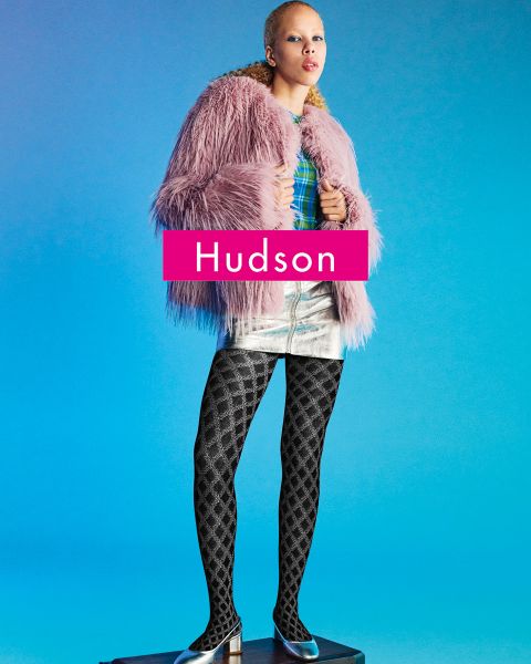 Una persona está de pie en un pedestal frente a un fondo azul, vistiendo un abrigo de piel sintética rosa, una blusa a cuadros, una falda metálica plateada, medias negras estampadas y zapatos de punta. La palabra "Hudson" aparece en negrita a través de la imagen.