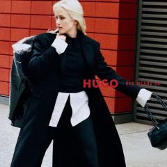 Una mujer con un abrigo negro y una camisa blanca posa junto al letrero "HUGO RED MEANS GO".
