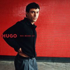 Un hombre con una chaqueta de punto negra y pantalones grises está de pie frente a una pared de azulejos rojos con las palabras "HUGO RED MEANS GO".
