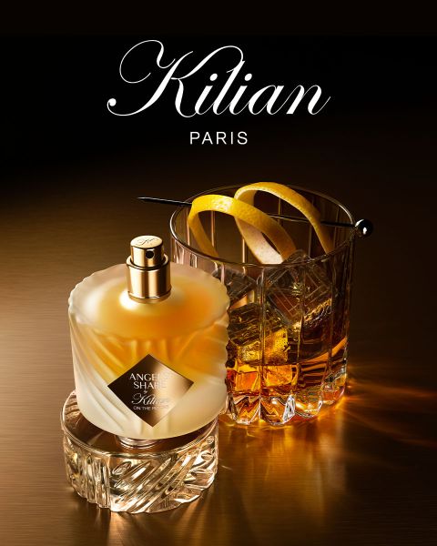 Un frasco del perfume Kilian Paris Angels' Share está junto a un vaso de whisky con hielo y una guarnición, ambos sobre una superficie marrón reflectante con iluminación suave. Arriba se ve el logo de Kilian Paris.