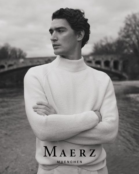 Un hombre con un jersey blanco de punto está de pie con los brazos cruzados junto a un río, con un puente de piedra arqueado y árboles desnudos al fondo. El texto "MAERZ MUENCHEN" aparece en la parte inferior de la imagen en blanco y negro.