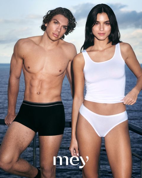 Un hombre en calzoncillos bóxer negros y una mujer en una camiseta sin mangas blanca y ropa interior están de pie uno al lado del otro al aire libre junto a una barandilla, con el mar y el cielo nublado de fondo. La palabra "mey" está en la parte inferior.