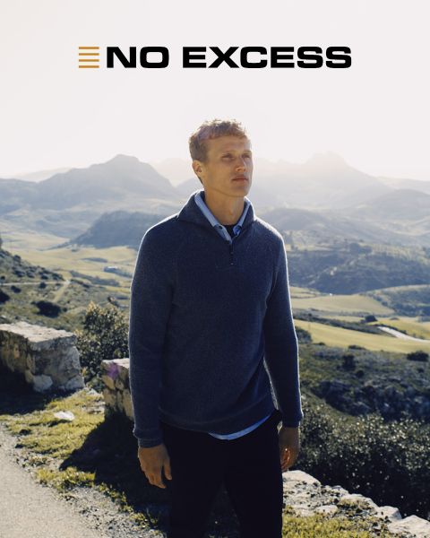 Un hombre con un suéter azul está de pie al aire libre en un día soleado, con montañas y valles al fondo. Las palabras "NO EXCESS" aparecen en negrita en la parte superior de la imagen.