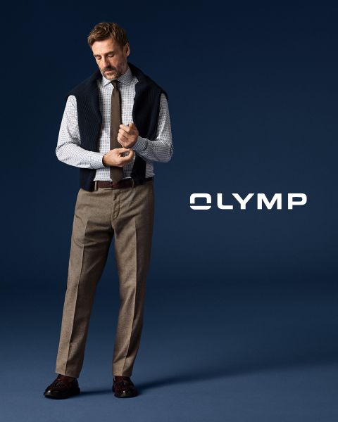 Un hombre con pantalones de traje, camisa a cuadros y corbata con un jersey drapeado sobre los hombros está de pie frente a un fondo azul oscuro, junto al cual se puede leer la palabra "OLYMP".