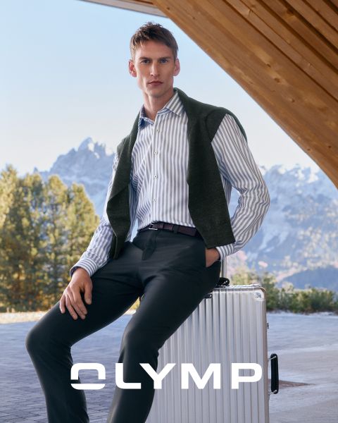 Un hombre con una camisa a rayas y pantalones oscuros está sentado en una maleta plateada al aire libre, con un suéter sobre los hombros. En el fondo se ven montañas nevadas y árboles. En la parte inferior está la palabra "OLYMP".