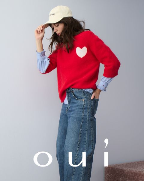 Una mujer que lleva un jersey rojo con un corazón blanco, vaqueros azules y un gorro blanco posa delante de una pared clara. En la parte inferior de la imagen está la palabra "oui" en letras blancas.