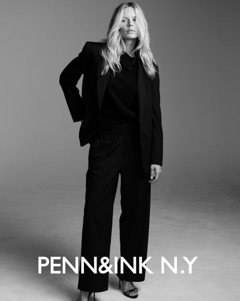 Una mujer con cabello largo y ondulado lleva un traje oscuro y sandalias de tacón alto, y está de pie con confianza en un estudio con un fondo de un solo color. El texto en la parte inferior dice "PENN&INK N.Y."
