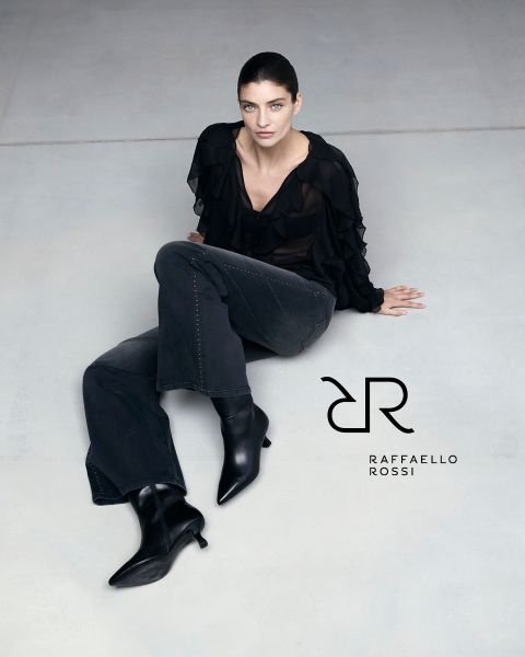 Una mujer de cabello oscuro está sentada en un suelo gris claro, vistiendo una blusa negra con volantes, jeans oscuros y botas negras puntiagudas. A su lado se puede ver el logo de Raffaello Rossi.