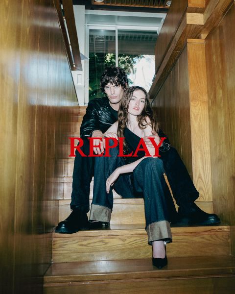 Un hombre y una mujer, vestidos con ropa oscura y elegante, están sentados en una escalera de madera en un interior. La palabra "REPLAY" aparece prominentemente en el centro de la imagen en letras mayúsculas rojas.