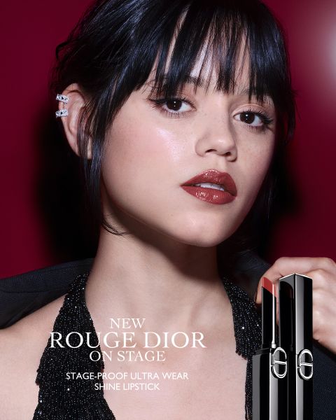 Una mujer con un labial rojo oscuro brillante posa frente a un fondo rojo intenso, luciendo un top halter negro brillante y orejeras. Sostiene un tubo de Rouge Dior Shine Lipstick. El texto promociona el nuevo labial de ultra duración a prueba de escenarios.