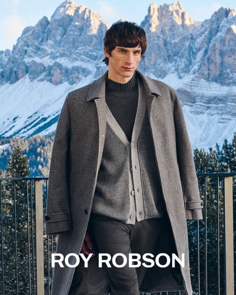Un hombre está de pie al aire libre frente a montañas nevadas y lleva un abrigo de pata de gallo gris, un jersey gris y pantalones grises. El texto "ROY ROBSON" aparece en la parte inferior de la imagen.