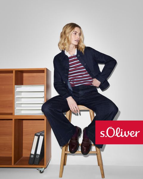 Una mujer con chaqueta y pantalones vaqueros oscuros, que lleva una camiseta de rayas, está sentada en un taburete de madera junto a una estantería de madera con archivos. El logotipo de s.Oliver aparece en la esquina inferior derecha.