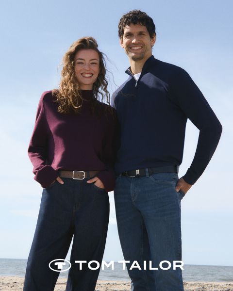 Una mujer sonriente con un jersey color castaño y un hombre con un jersey azul marino están de pie uno al lado del otro en una playa. Ambos visten vaqueros. El cielo está despejado y azul. El logo "Tom Tailor" aparece en la parte inferior.