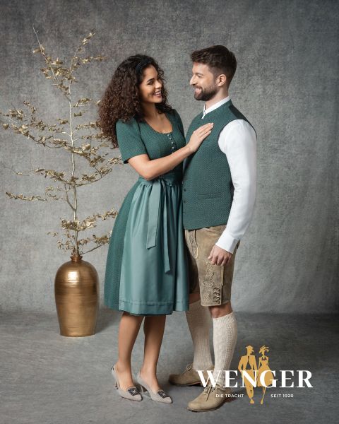 Una mujer sonriente con un dirndl verde está de pie junto a un hombre vestido con ropa tradicional bávara, que incluye pantalones de cuero y un chaleco. Están uno frente al otro delante de un fondo de estudio con un jarrón dorado y una rama decorativa. Logotipo de "Wenger" en la parte inferior.