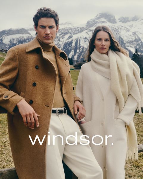 Un hombre con un abrigo marrón y una mujer con un abrigo y una bufanda color crema están de pie al aire libre con montañas al fondo. La palabra "windsor." se puede leer en la parte inferior de la imagen.