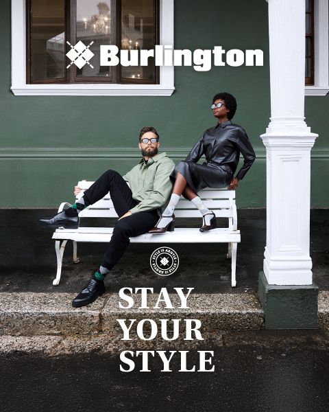 Un hombre y una mujer con atuendos elegantes posan con confianza en un banco blanco frente a una pared verde con los textos "Burlington" y "Stay Your Style" encima y debajo de ellos.