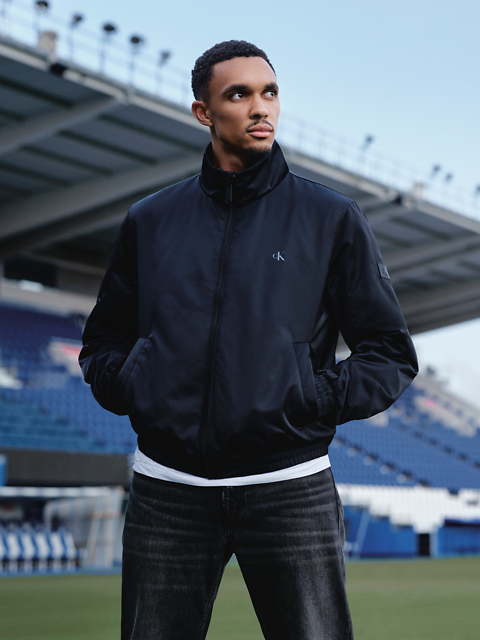 Un hombre con una chaqueta negra y vaqueros está de pie en un estadio y mira hacia un lado con las manos en los bolsillos. En el fondo se ven asientos de estadio y un gran edificio.