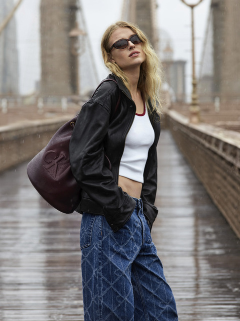 Una mujer con gafas de sol, chaqueta negra, top corto blanco y vaqueros azules anchos está de pie en un puente lluvioso y lleva un bolso grande marrón castaño al hombro. El puente y las lámparas están borrosos al fondo.