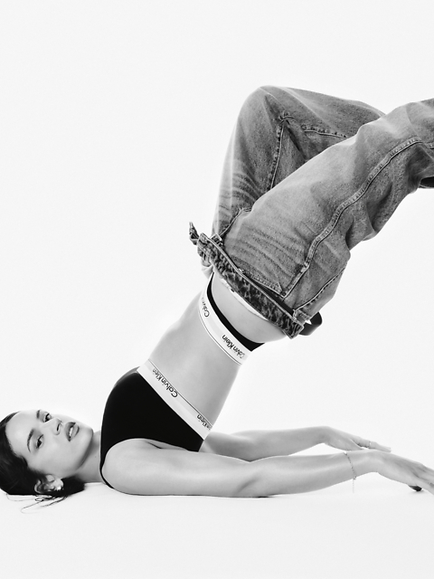 Fotografía en blanco y negro de una mujer con un sujetador deportivo de Calvin Klein y jeans, acostada boca arriba con las piernas dobladas sobre la cabeza, los brazos extendidos en el suelo y mirando a la cámara.