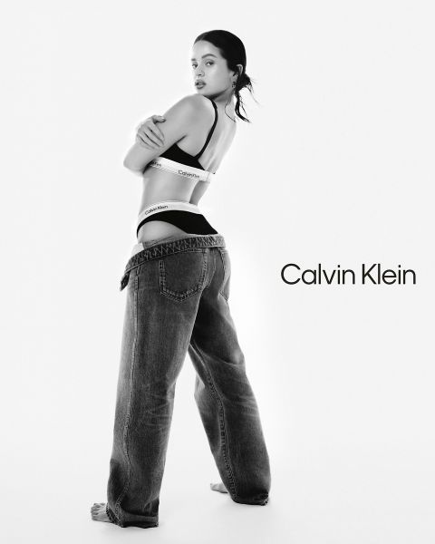 Una mujer posa en ropa interior Calvin Klein y vaqueros anchos, mirando por encima del hombro. El fondo es blanco liso con el logo de Calvin Klein en letras negras en el lado derecho. La imagen está en blanco y negro.