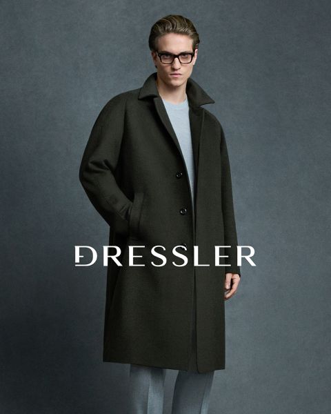 Un hombre que lleva gafas, un abrigo oscuro y pantalones gris claro está de pie frente a un fondo gris. La palabra "DRESSLER" está impresa en grandes letras blancas a través de la imagen.