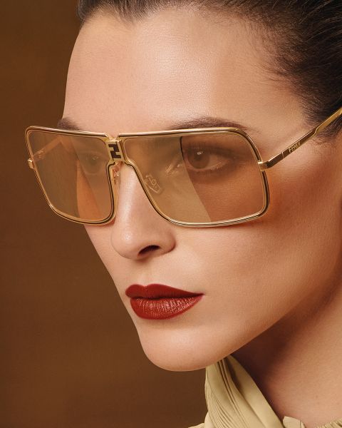 Primer plano de una persona que lleva unas gafas de sol grandes, cuadradas, con montura dorada y cristales marrones. Tiene la piel lisa, el pelo peinado hacia atrás, lleva un labial rojo intenso y una blusa de color claro sobre un fondo marrón.