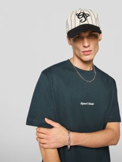 Un joven que lleva una camiseta negra con la inscripción "Original Studio", un collar plateado, una pulsera plateada y una gorra de béisbol a rayas color crema y negro, está de pie frente a un fondo liso de color gris claro.