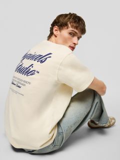 Un joven con cabello castaño corto, que lleva una camiseta color crema con un estampado azul marino en la espalda, jeans azul claro y zapatillas de deporte color beige, está sentado en el suelo mirando por encima del hombro a la cámara.
