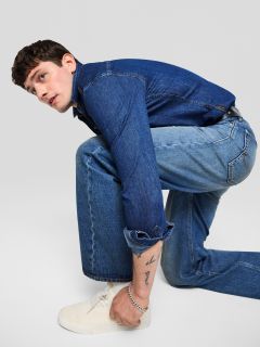 Un joven con chaqueta y pantalones vaqueros está arrodillado y mirando hacia un lado. Lleva zapatillas blancas y tiene un tatuaje visible en el antebrazo. El fondo es de color gris claro uniforme.