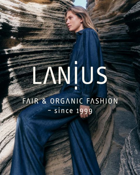 Una mujer con un vestido largo azul oscuro está sentada entre formaciones rocosas en capas. El texto superpuesto dice: "LANIUS FAIR & ORGANIC FASHION - desde 1999.