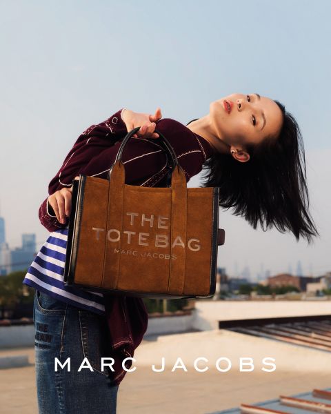 Una mujer posa al aire libre con un bolso marrón de Marc Jacobs en la mano, se inclina hacia atrás con la cabeza ladeada y lleva una chaqueta color castaño y vaqueros azules. En la imagen aparecen las palabras "EL BOLSO TOTE MARC JACOBS" y "MARC JACOBS".