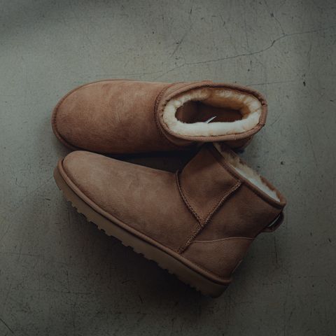 Un par de botas de invierno de ante marrón claro con un forro grueso de felpa clara, sobre un suelo de hormigón gris ligeramente agrietado.