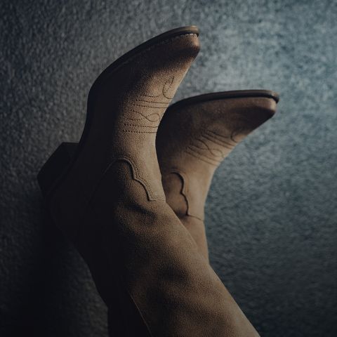 Primer plano de un par de botas vaqueras marrones de ante con costuras decorativas sobre un fondo oscuro y texturizado. Las botas están orientadas hacia arriba, destacando su diseño y material.