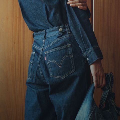 Una persona con una camisa vaquera oscura y vaqueros está de pie frente a una pared de madera, sosteniendo una chaqueta vaquera en una mano mientras da la espalda a la cámara.