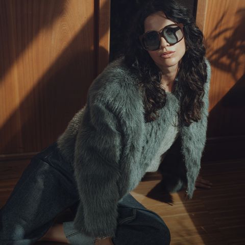 Una mujer con cabello oscuro ondulado, que lleva gafas de sol grandes, una chaqueta de piel gris y vaqueros oscuros, está sentada en un suelo de madera cerca de una pared de madera con una iluminación dramática.