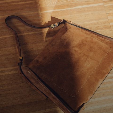 Un bolso bandolera de ante marrón con una correa ancha está sobre un suelo de madera iluminado por el sol, donde la luz y las sombras crean una apariencia cálida y texturizada.