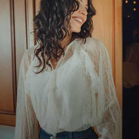 Una mujer con cabello oscuro ondulado, que lleva una blusa blanca de encaje y jeans azules, está de pie sonriendo en una habitación con paneles de madera e iluminación cálida, mirando hacia un lado.