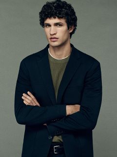 Un hombre con cabello oscuro y rizado, que lleva un blazer negro sobre un jersey de punto verde, está de pie con los brazos cruzados frente a un fondo gris liso y mira al frente con una expresión neutral.