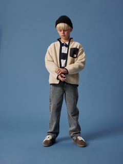 Un chico con cabello rubio lleva una gorra negra, una chaqueta de forro polar color crema sobre una camisa a rayas, vaqueros grises y zapatillas de deporte. Está de pie frente a un fondo azul liso y mira hacia abajo con las manos juntas.