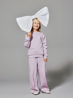 Una joven sonriente con un chándal color lavanda de pie frente a un fondo gris, con un lazo blanco muy grande en la cabeza.