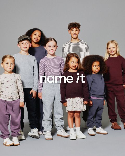 Un grupo de ocho niños diferentes están juntos frente a un fondo gris, todos vestidos con atuendos casuales a juego en colores neutros y pastel. El texto "name it" aparece en el centro de la imagen.