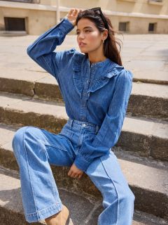 Mujer sentada en una escalera de piedra al aire libre, vestida con una blusa vaquera de manga larga con detalles de volantes y unos vaqueros a juego de talle alto. Tiene el pelo oscuro, lleva gafas de sol en la cabeza y mira pensativa a la distancia.