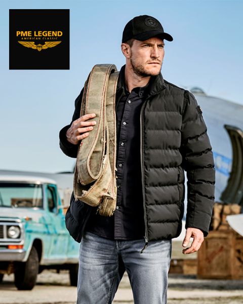 Un hombre con una chaqueta acolchada negra, gorro y vaqueros lleva una correa de lona enrollada sobre el hombro y está de pie al aire libre cerca de un camión azul antiguo y un avión. El logotipo de PME Legend aparece en la esquina superior izquierda.