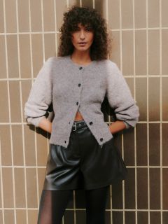 Una mujer con cabello rizado está de pie junto a una pared de azulejos. Lleva un cárdigan de punto gris claro y esponjoso, pantalones cortos de cuero negros de talle alto y medias negras, con las manos en los bolsillos y una expresión facial segura.