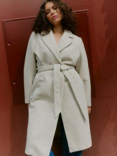 Una persona con cabello rizado y un cinturón beige claro está de pie frente a una pared de color marrón rojizo y mira ligeramente hacia abajo con una expresión relajada.